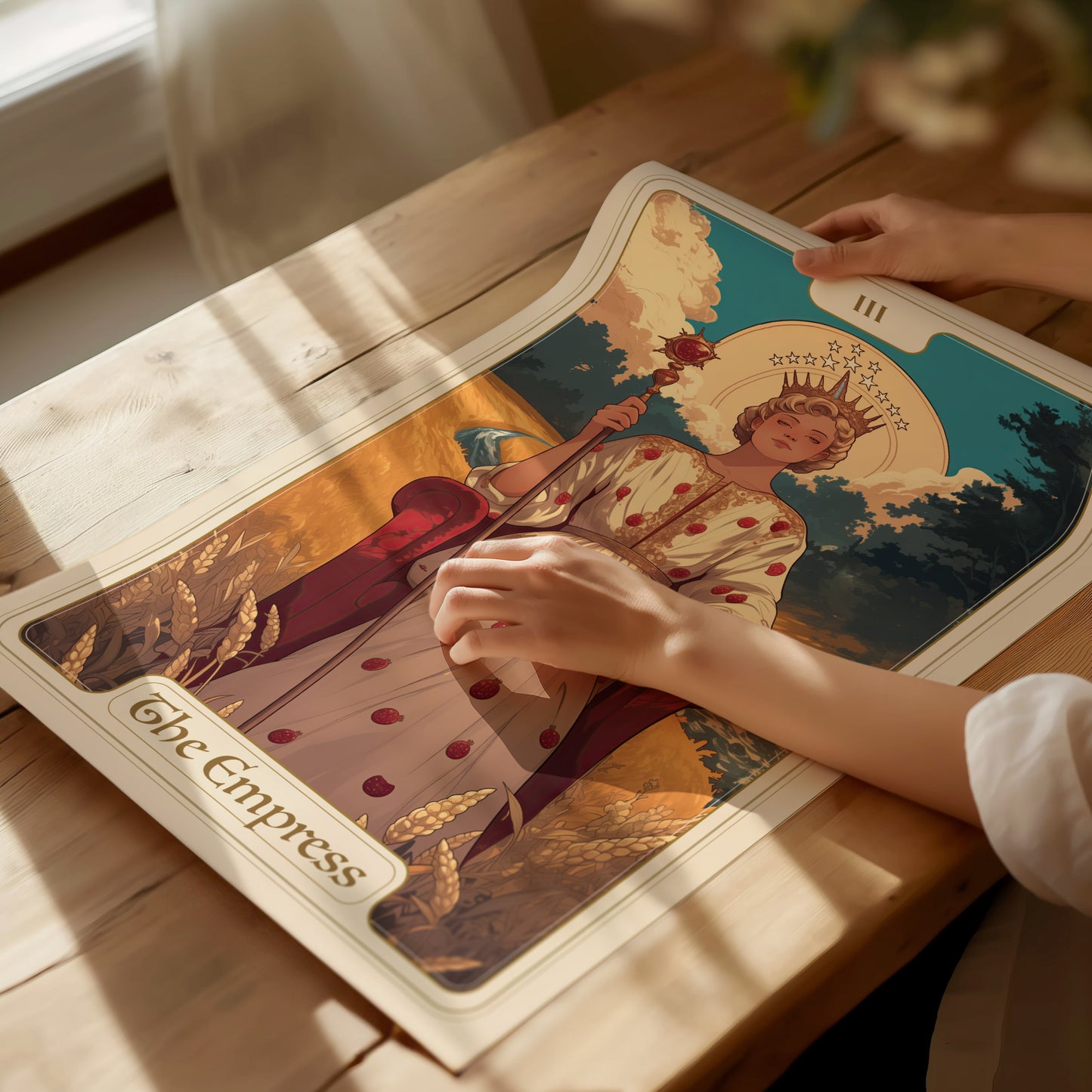 A person gently unrolls an Empress Tarot poster on a wooden table, featuring a regal figure with a crown and wheat motifs, surrounded by warm sunlight.