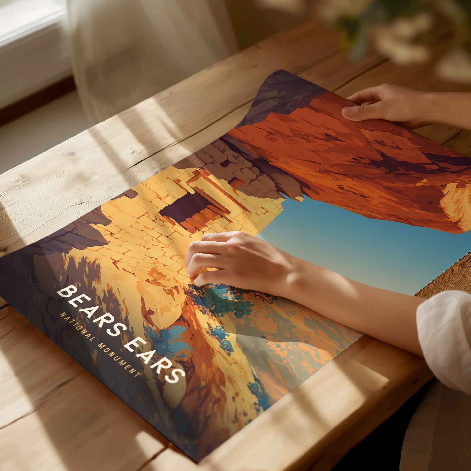 A person examines a vibrant poster of Bears Ears National Monument, depicting red sandstone cliffs and ancient ruins, on a wooden table with soft natural light streaming.