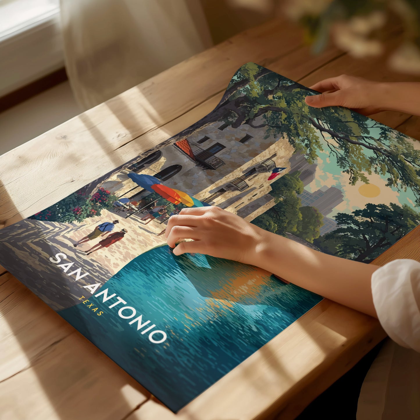 A person holds a vibrant poster featuring a colorful depiction of San Antonio, Texas, with a historic building and a river scene, placed on a wooden table.