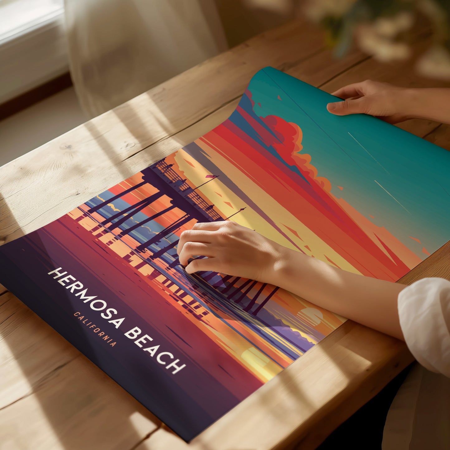A person adjusts a vibrant poster on a wooden table, depicting a colorful sunset at Hermosa Beach Pier, California, with bold hues of orange, pink, and blue.