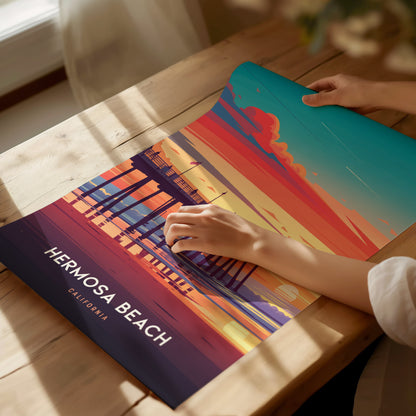 A person adjusts a vibrant poster on a wooden table, depicting a colorful sunset at Hermosa Beach Pier, California, with bold hues of orange, pink, and blue.