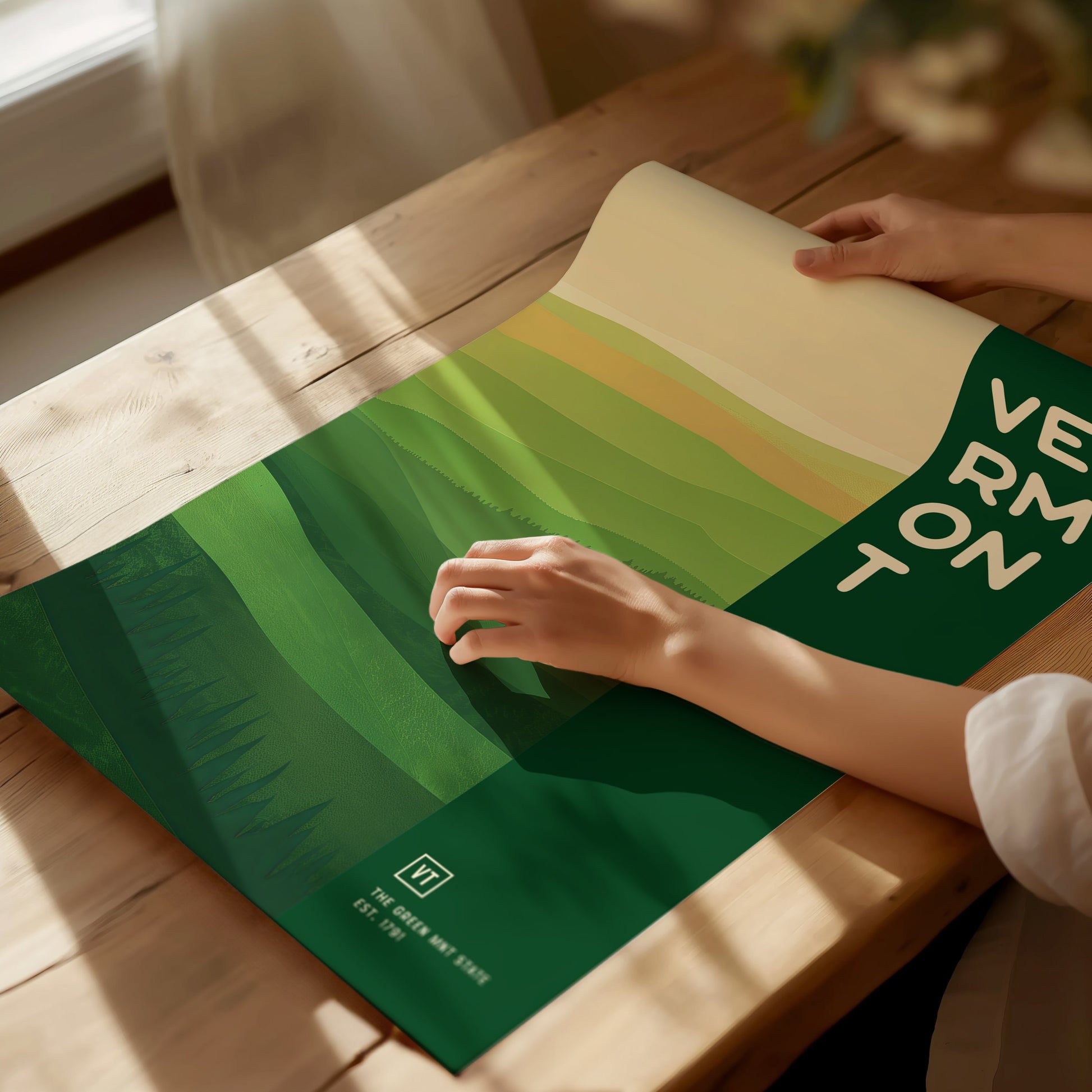 A person unrolls a Vermont-themed poster with green abstract landscape patterns on a wooden table. Sunlight illuminates the scene, highlighting the minimalist design and bold lettering.