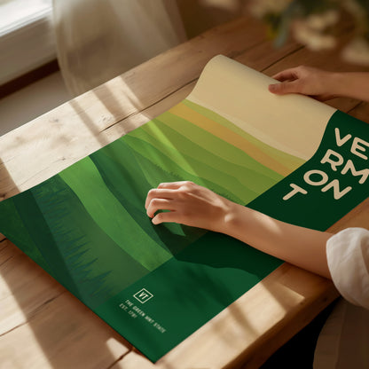 A person unrolls a Vermont-themed poster with green abstract landscape patterns on a wooden table. Sunlight illuminates the scene, highlighting the minimalist design and bold lettering.