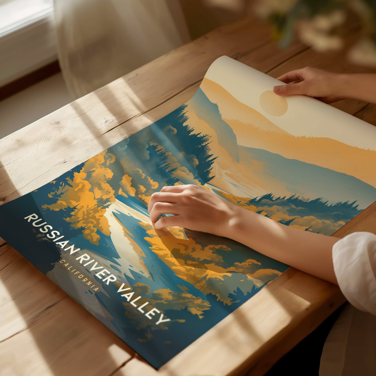 A person unrolls a Russian River Valley poster on a wooden table, featuring stylized landscapes with blue.
