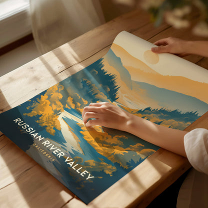 A person unrolls a Russian River Valley poster on a wooden table, featuring stylized landscapes with blue.