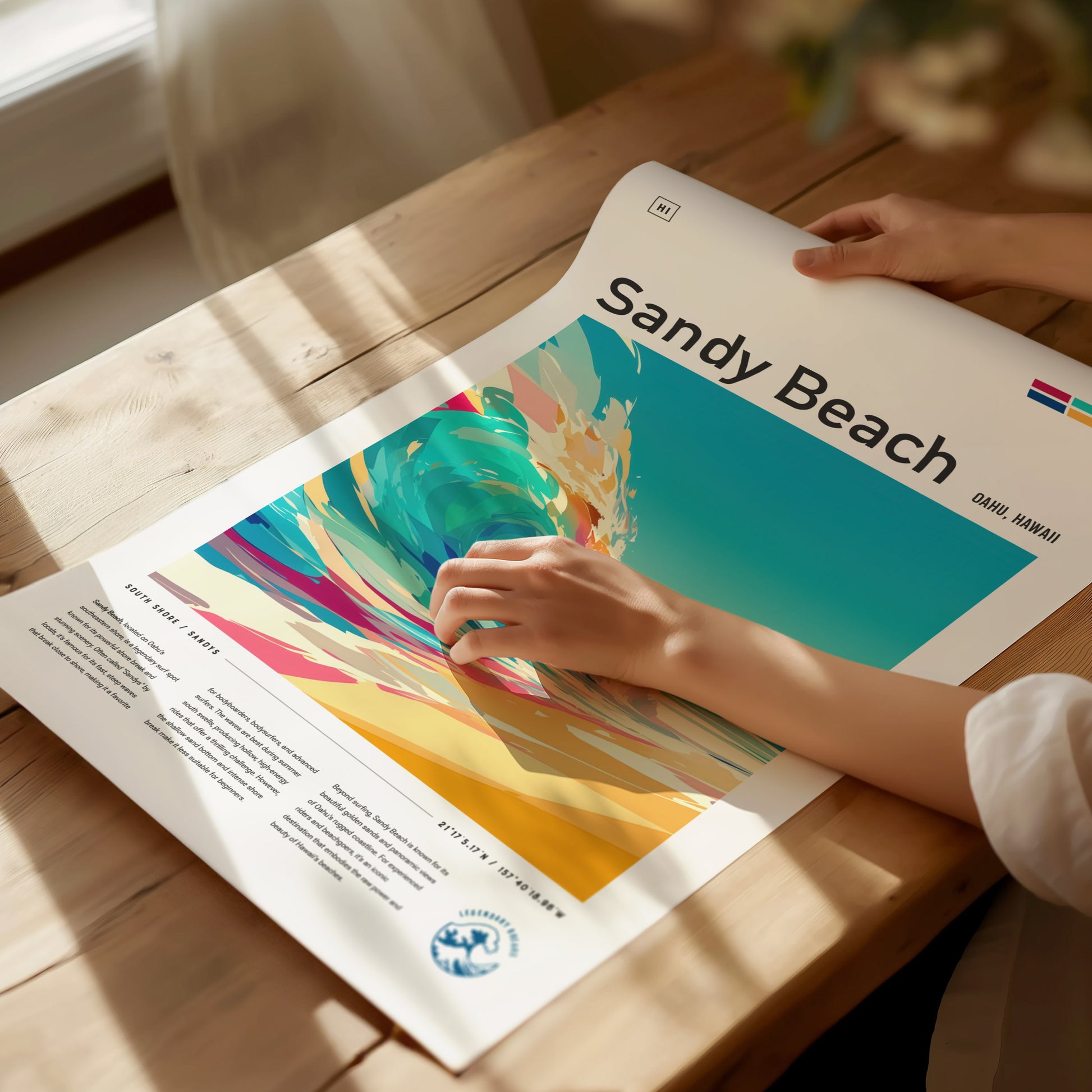 A person is holding a colorful Sandy Beach poster on a wooden table, featuring vibrant abstract waves and beach scenery. Sunlight casts soft shadows across the scene.