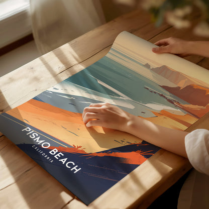 A person examines a vibrant Pismo Beach poster on a wooden table, featuring abstract coastal scenery with bold colors. Sunlight casts soft shadows on the tabletop.
