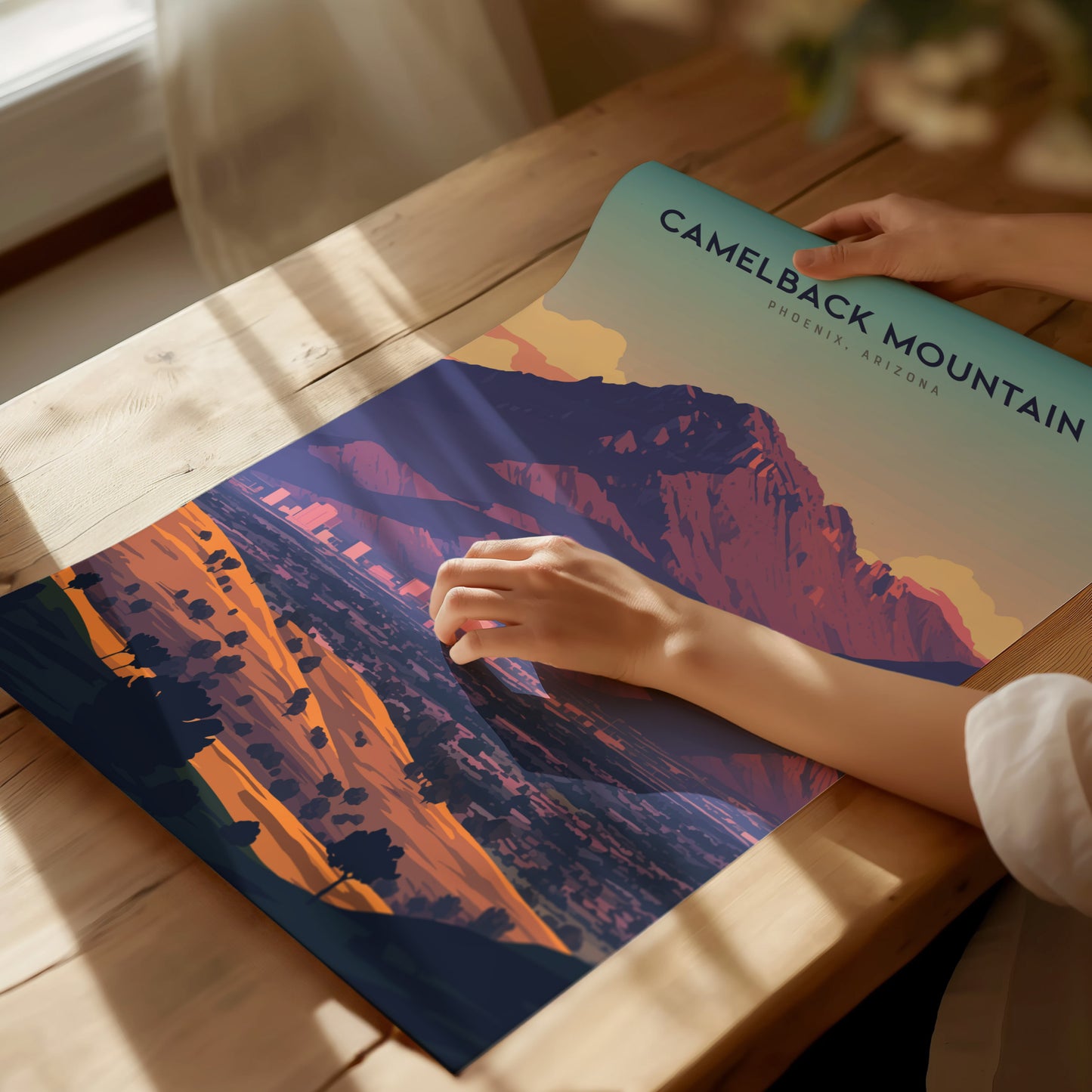 A person holds a vibrant Camelback Mountain poster showing the dramatic silhouette and rich desert tones. The poster lies on a wooden table, with sunlight streaming.