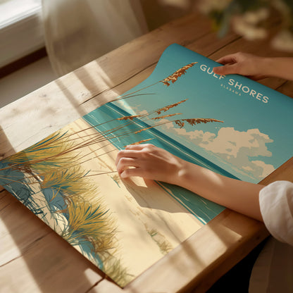 A person is holding a Gulf Shores, Alabama poster featuring a coastal scene with sandy beach, sea oats,.