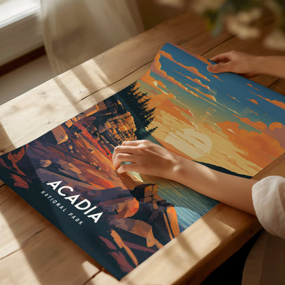 A person arranges an Acadia National Park poster on a wooden table, featuring vibrant cliffs and a colorful sunset. Sunlight streams through a nearby window, creating a serene atmosphere.