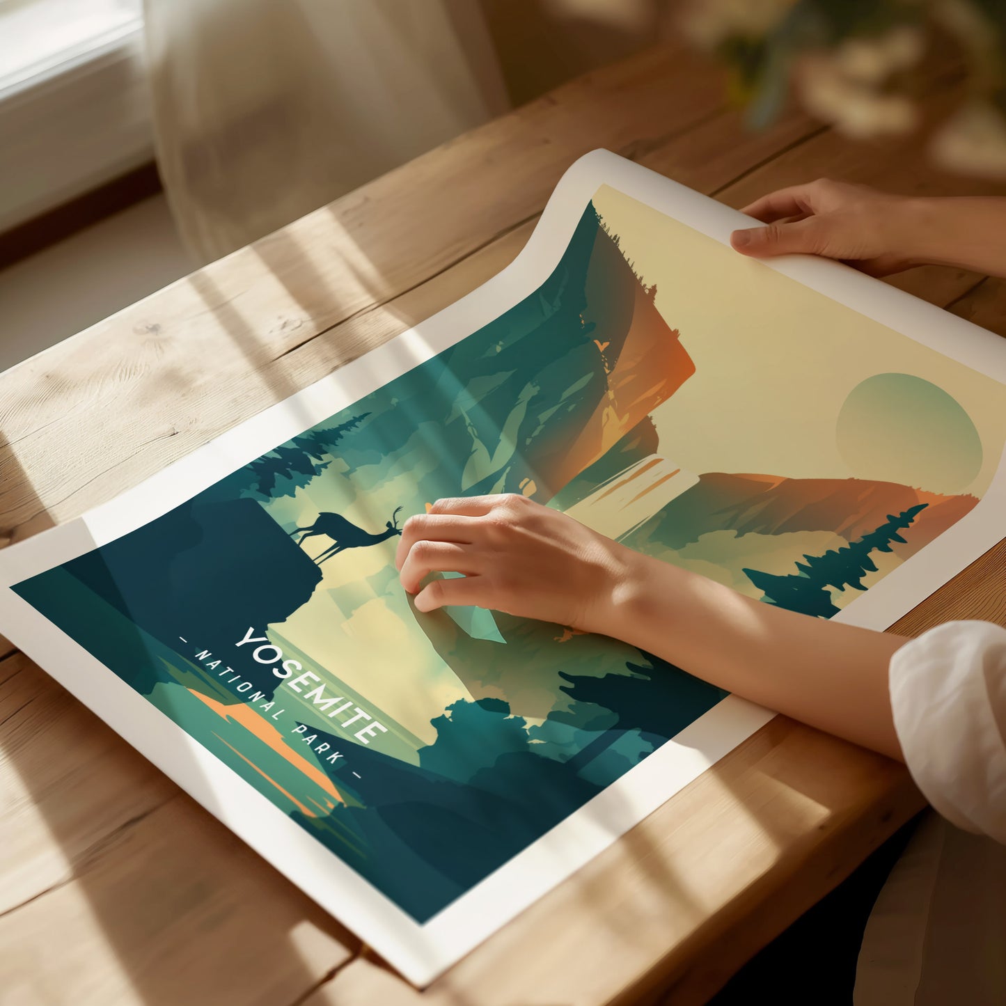 A person unrolls a minimalist Yosemite National Park poster on a wooden table, featuring stylized mountains, a deer silhouette,.
