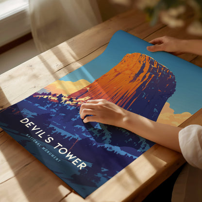 A person is holding a colorful poster of Devils Tower on a wooden table, with sunlight streaming in. The poster features the striking geological formation against a blue sky.
