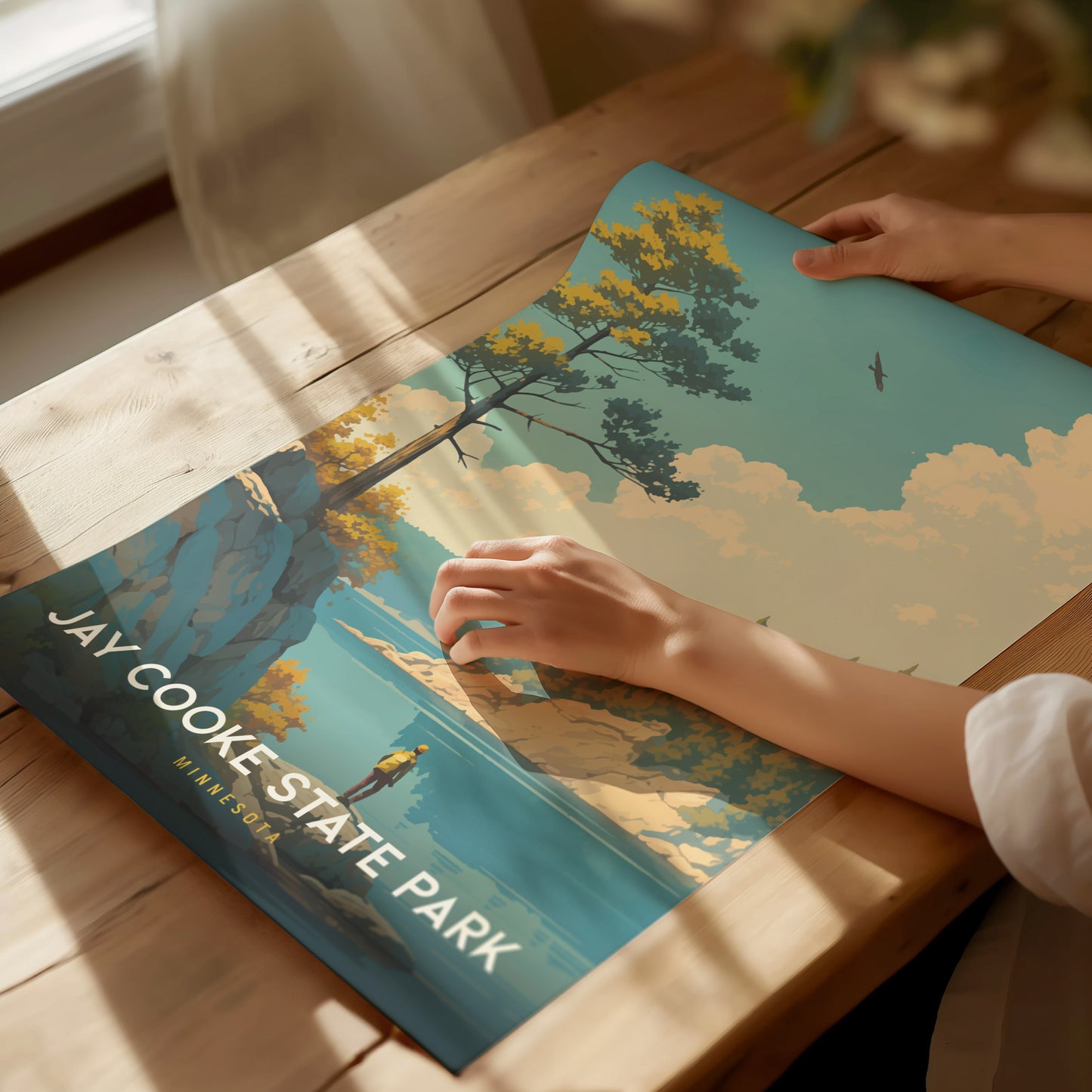 A person gently holds a colorful Jay Cooke State Park poster on a wooden table, featuring a scenic landscape with trees, clouds, and a bird in flight.