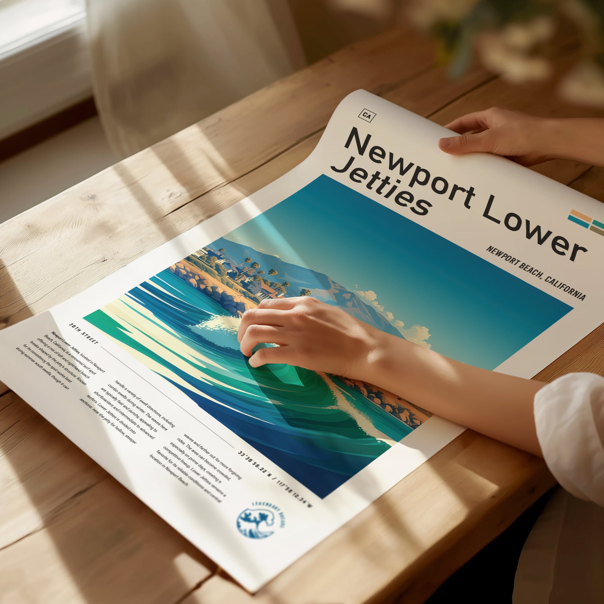 A person unrolls a vibrant Newport Lower Jetties poster on a wooden table, featuring a stylized beach scene with waves and palm trees under a clear blue sky.