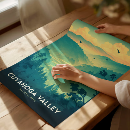 A person unrolls a vibrant Cuyahoga Valley National Park poster on a wooden table, featuring trees, hills, and birds against a colorful sky. Warm sunlight illuminates the scene.