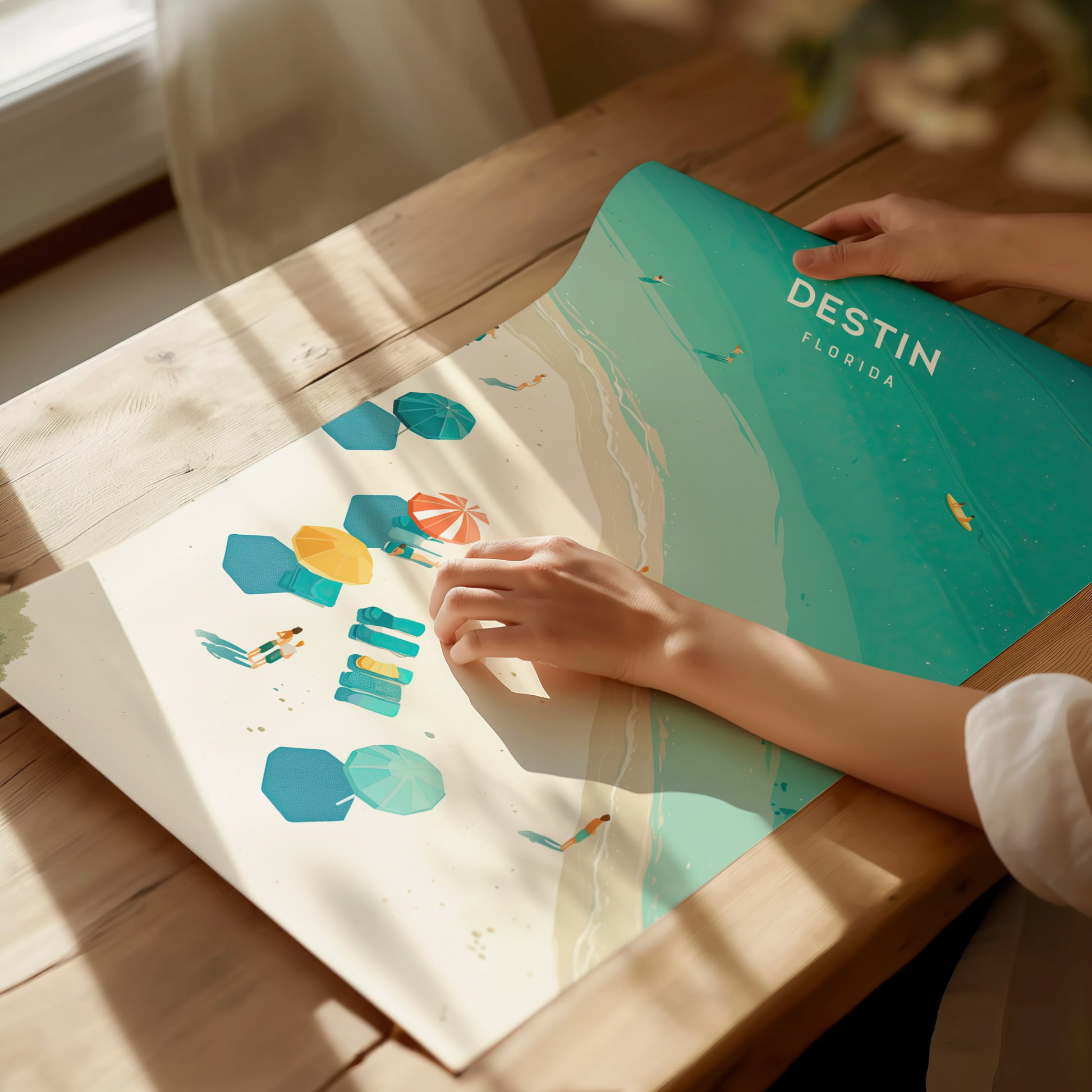 A person holds a vibrant Destin, Florida poster depicting colorful beach umbrellas on a sandy shore with turquoise waters. Sunlight casts gentle shadows on the wooden table.