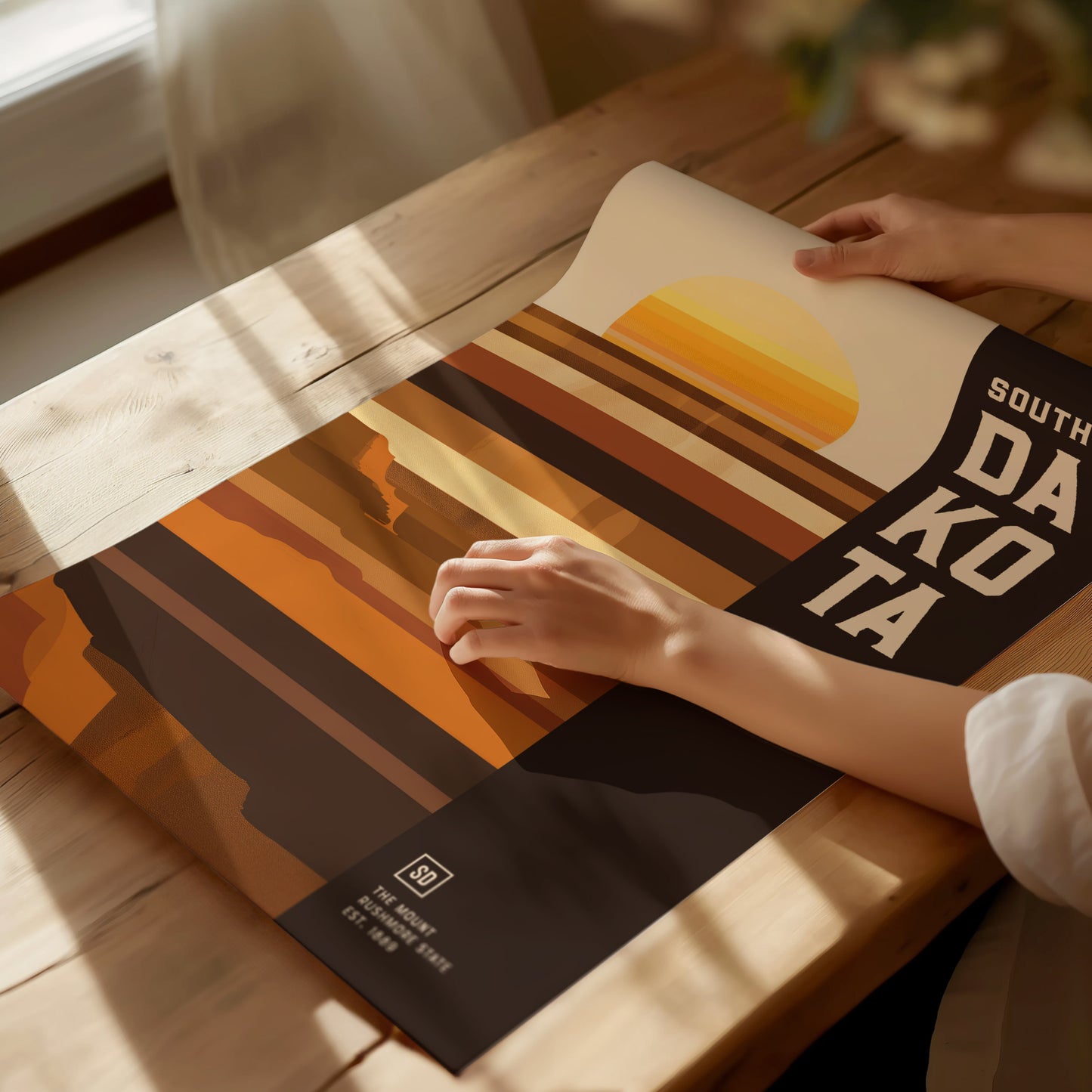 A person adjusts a South Dakota poster on a wooden table, featuring a minimalist geometric design with earthy tones and a stylized sunset. Sunlight streams through a nearby window.