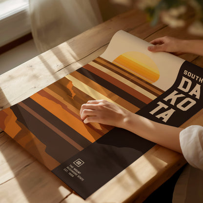 A person adjusts a South Dakota poster on a wooden table, featuring a minimalist geometric design with earthy tones and a stylized sunset. Sunlight streams through a nearby window.