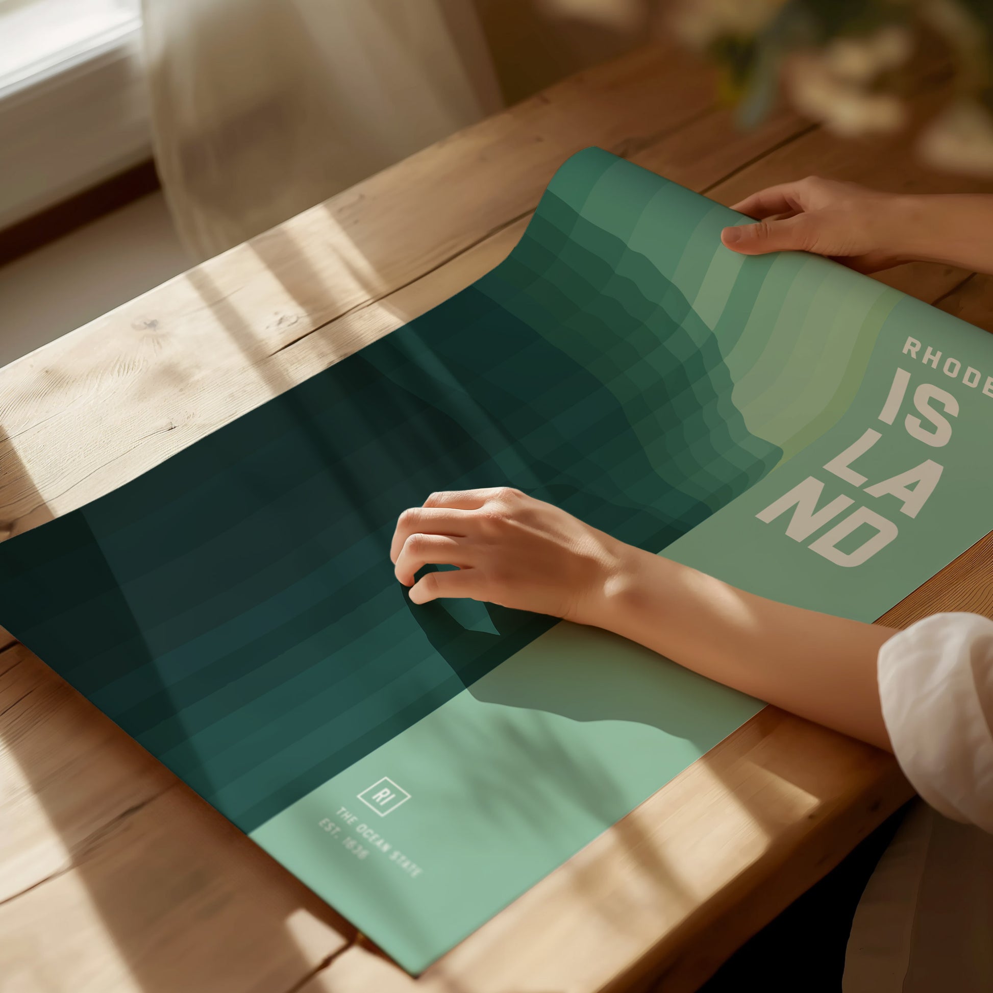 A person is smoothing out a green and blue abstract poster titled Rhode Island on a wooden table, with sunlight streaming in. The design features layered geometric shapes.