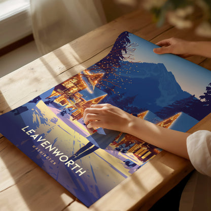 A person unrolls a vibrant Leavenworth, Washington poster on a wooden table, depicting a festive Bavarian village with illuminated buildings and mountain backdrop. Sunlight filters through a nearby window.