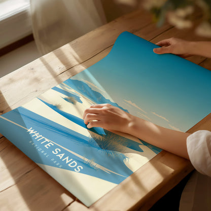 A person unrolls a vibrant White Sands National Park poster on a wooden table, showcasing blue skies and white sand dunes. Sunlight streams through a nearby window.