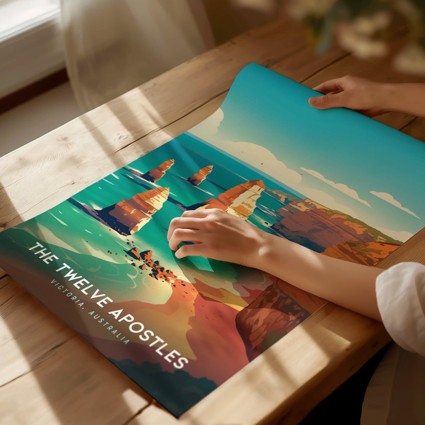 A person unrolls a vibrant poster of the Twelve Apostles on a wooden table, displaying the iconic limestone stacks against a turquoise ocean. Sunlight streams through a nearby window.