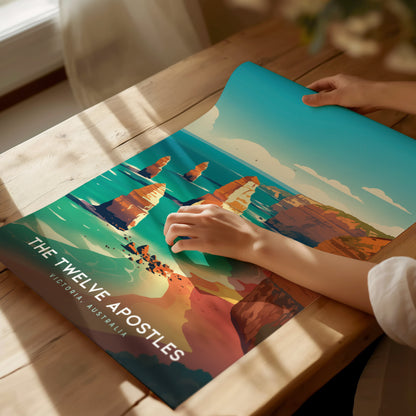 A person unrolls a vibrant poster of the Twelve Apostles on a wooden table, displaying the iconic limestone stacks against a turquoise ocean. Sunlight streams through a nearby window.