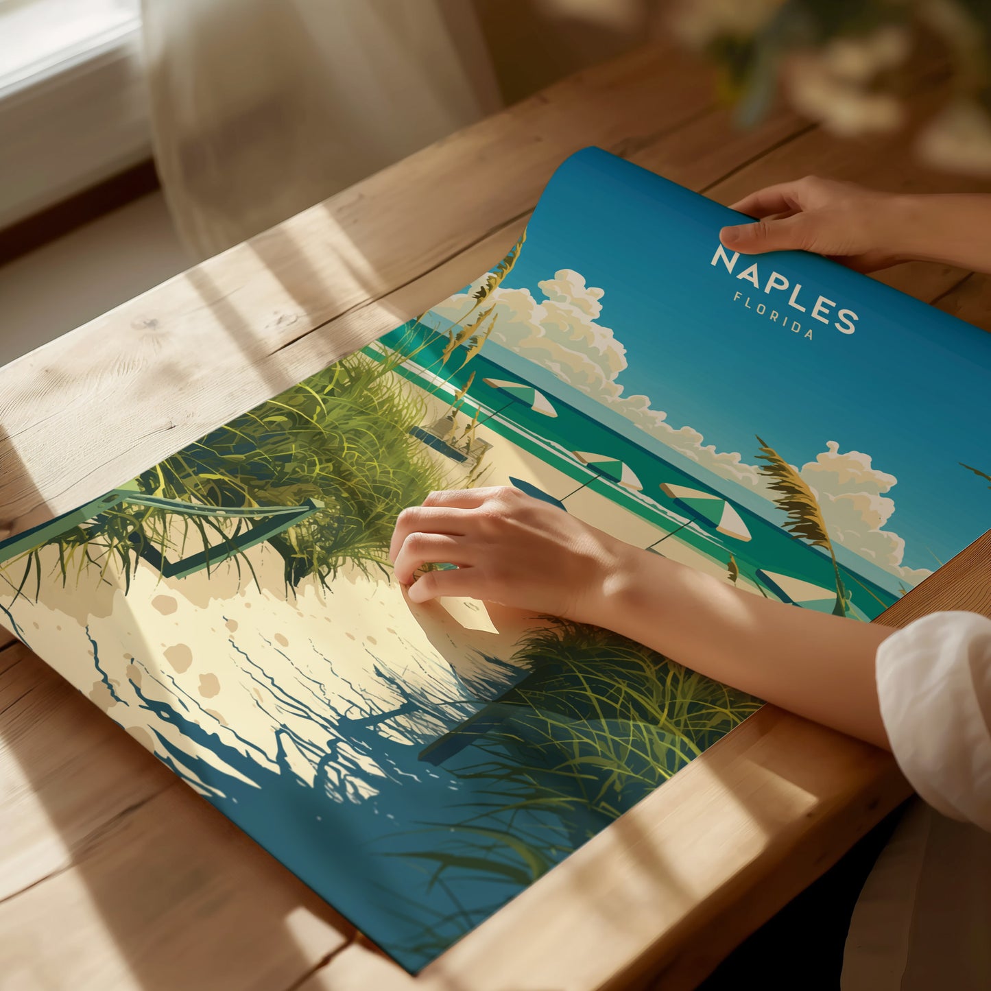 A person gently holds a vibrant poster depicting a beach scene with palm trees, sailboats, and clouds under a blue sky, labeled Naples Florida, on a sunlit wooden table.