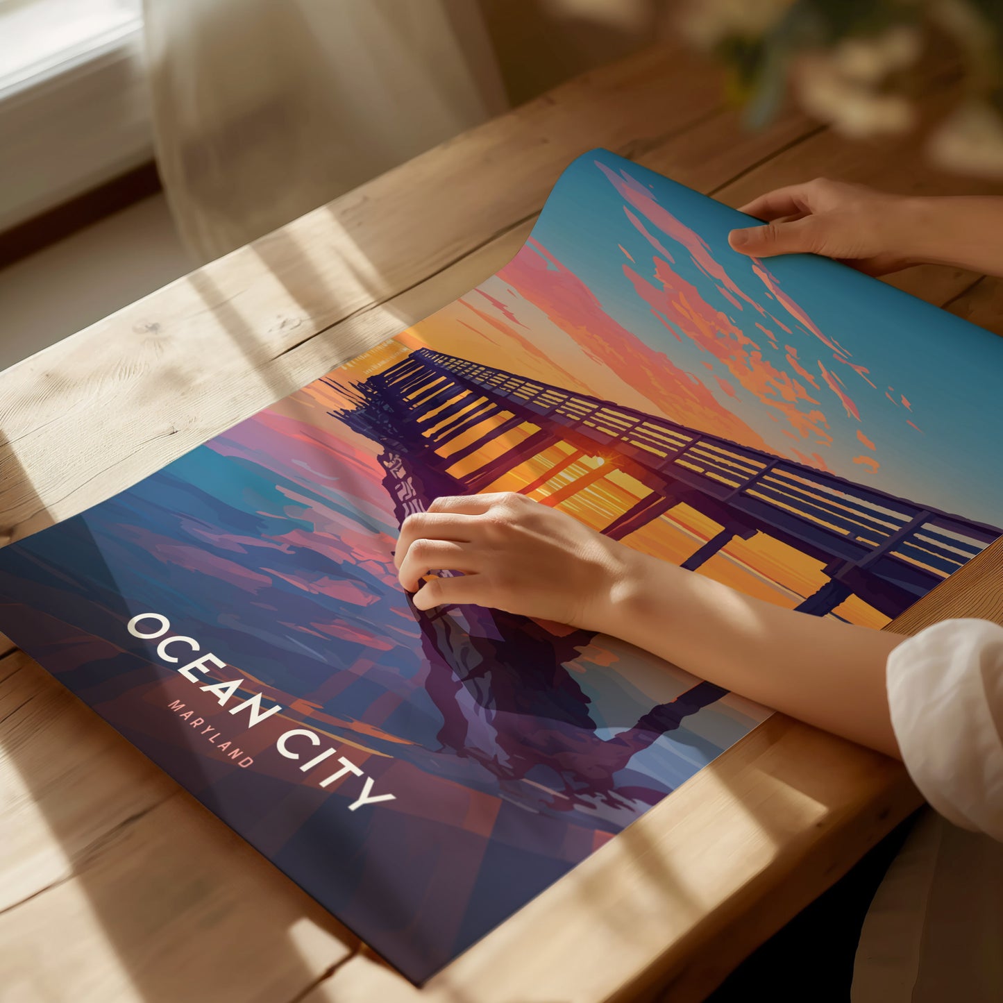 A person unrolls a vibrant Ocean City Maryland poster featuring a boardwalk pier at sunset, with pink and orange clouds against a blue sky, on a wooden table.