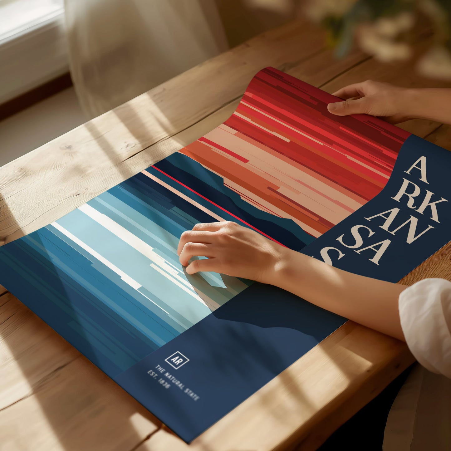 A person adjusts a vibrant, abstract Arkansas poster on a wooden table, featuring bold red and blue stripes with large white text. Sunlight casts soft shadows across the scene.