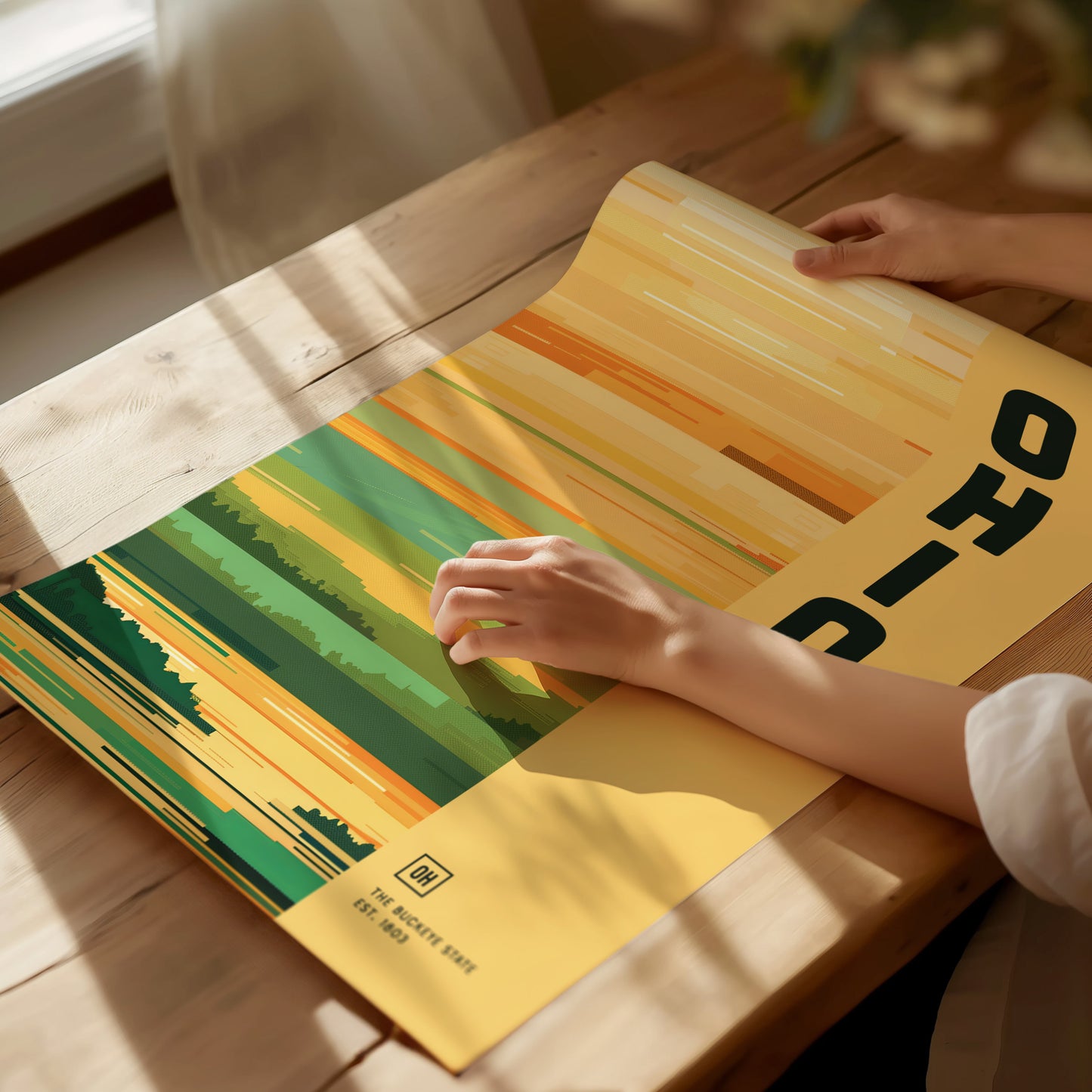 A person gently holds an Ohio poster with abstract green and yellow stripes on a wooden table, illuminated by soft natural light.