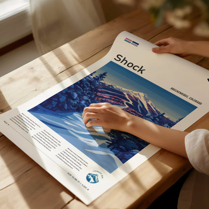 A person adjusts a colorful ski art poster titled Shock on a wooden table, depicting snow-covered trees and mountains in Breckenridge, Colorado, under natural lighting.