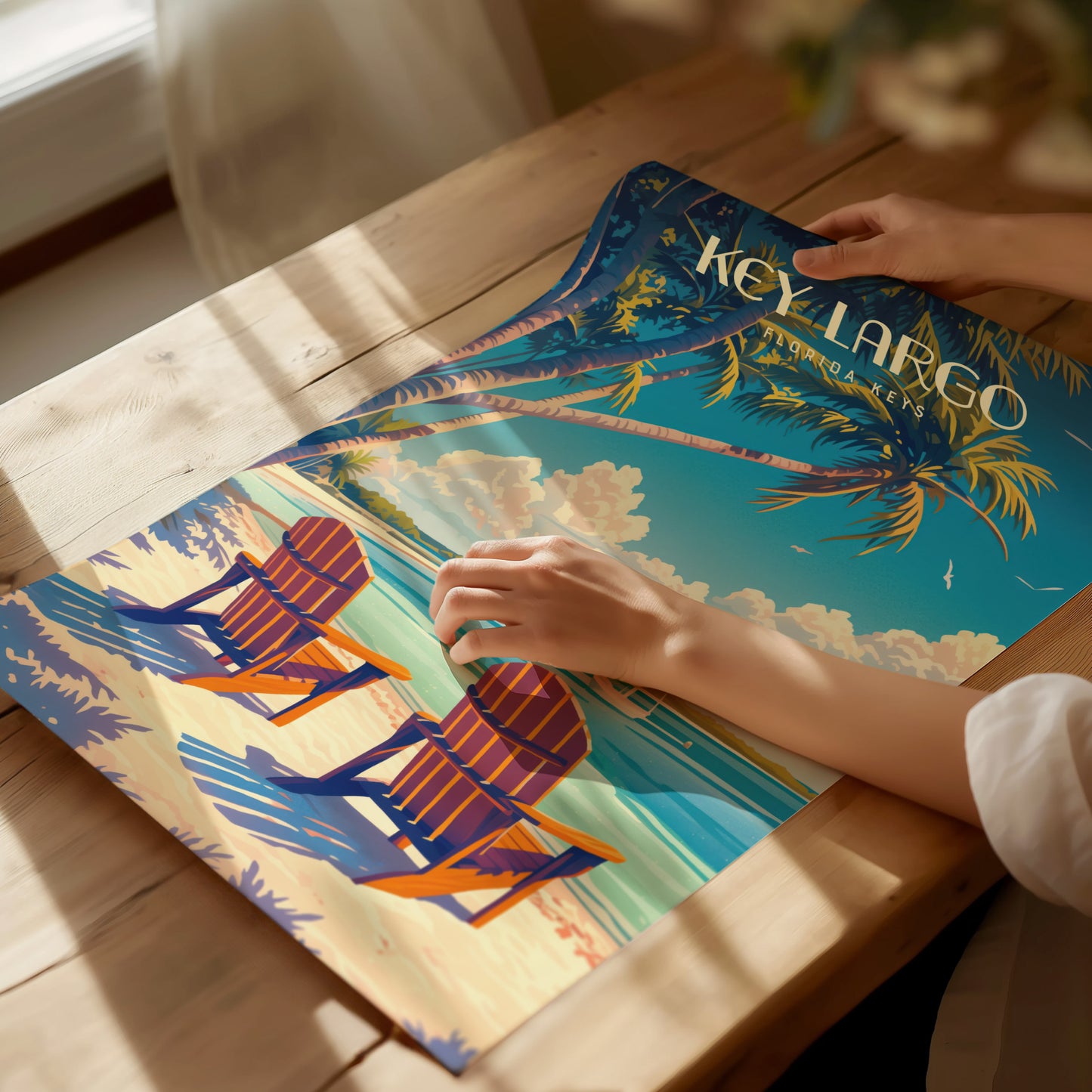 A person arranges a vibrant Key Largo poster featuring palm trees, Adirondack chairs, and a beach scene on a wooden table under warm.