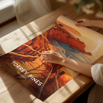 A person gently holds a vibrant Torrey Pines poster on a wooden table, featuring orange and blue coastal scenery with the text Torrey Pines, California prominently displayed.