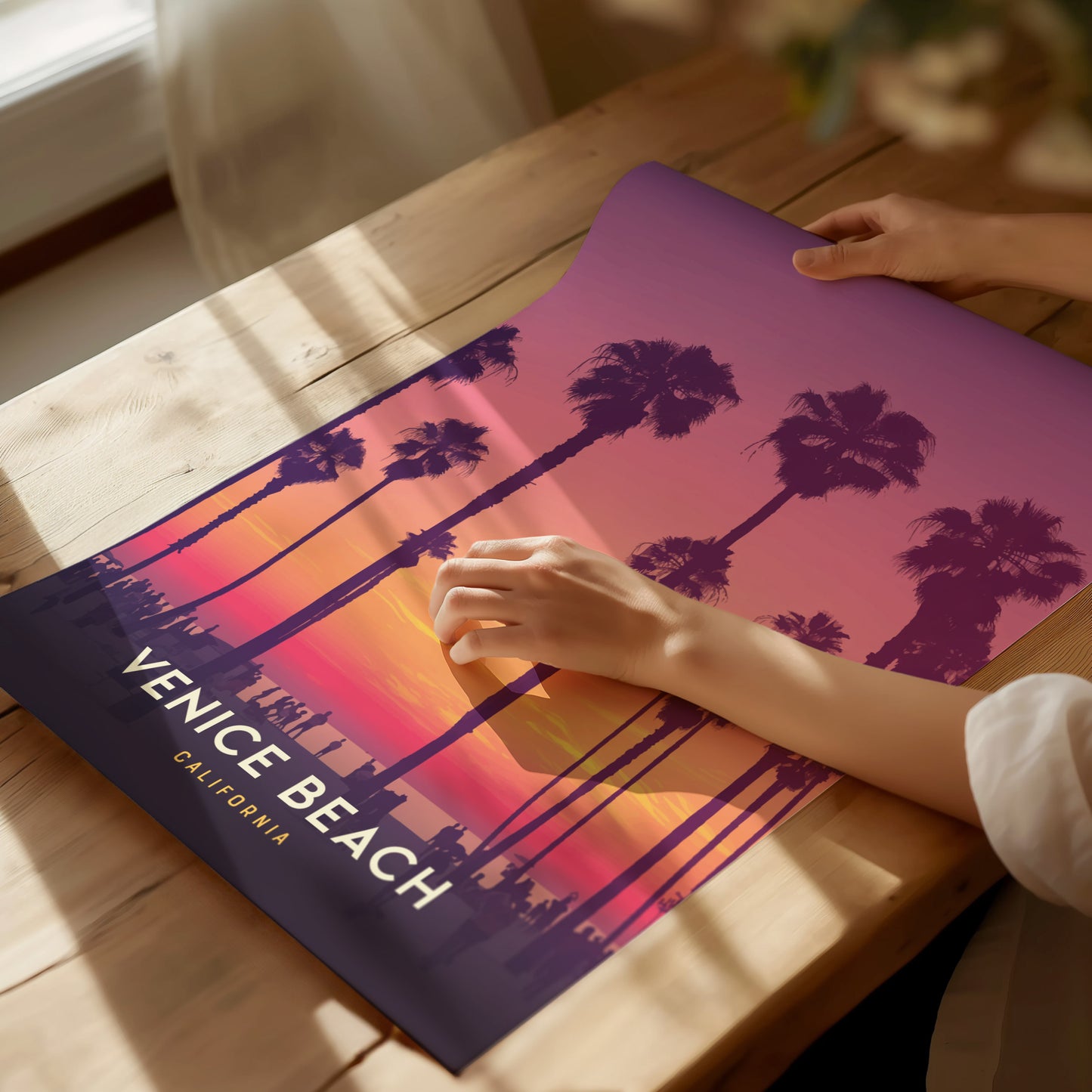 A person holds a Venice Beach poster with palm trees silhouetted against a vibrant purple and orange sunset. The poster rests on a wooden table in soft lighting.