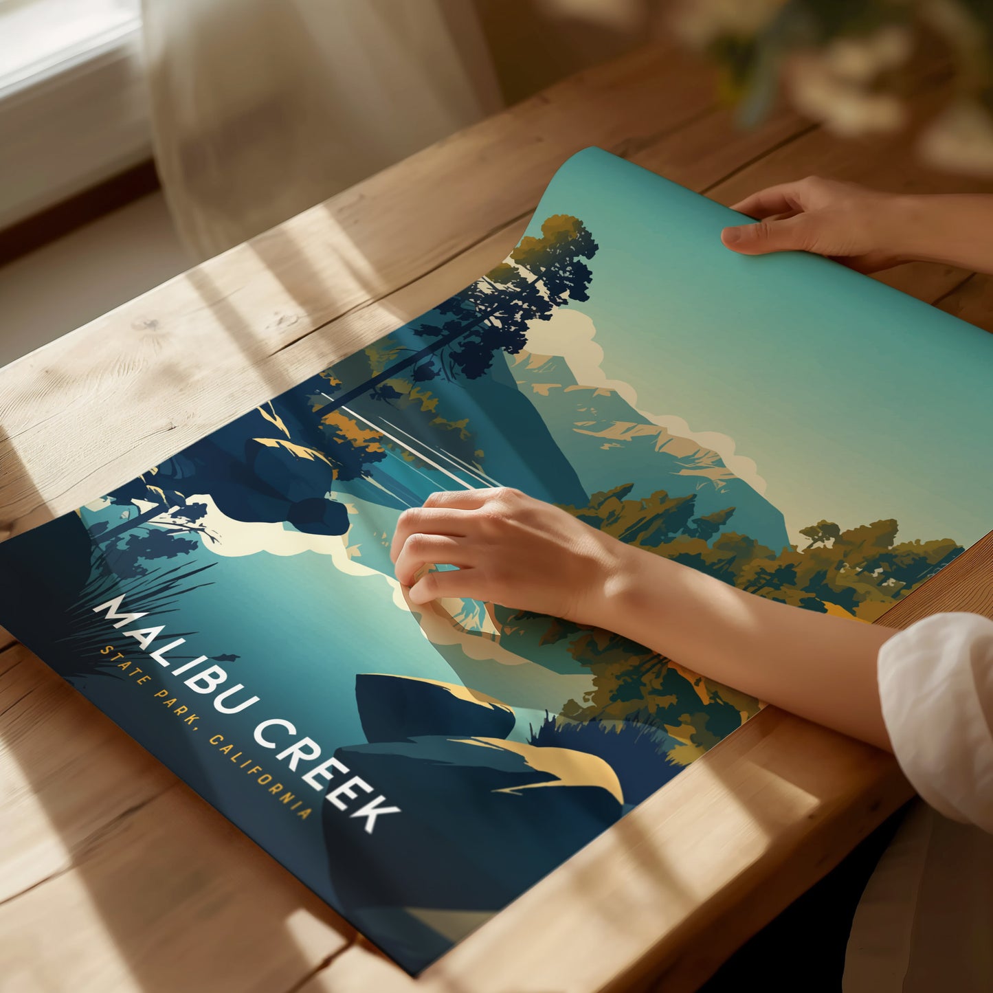 A person examines a colorful poster featuring Malibu Creek State Park on a wooden table, showcasing stylized landscapes with trees and mountains under bright lighting.