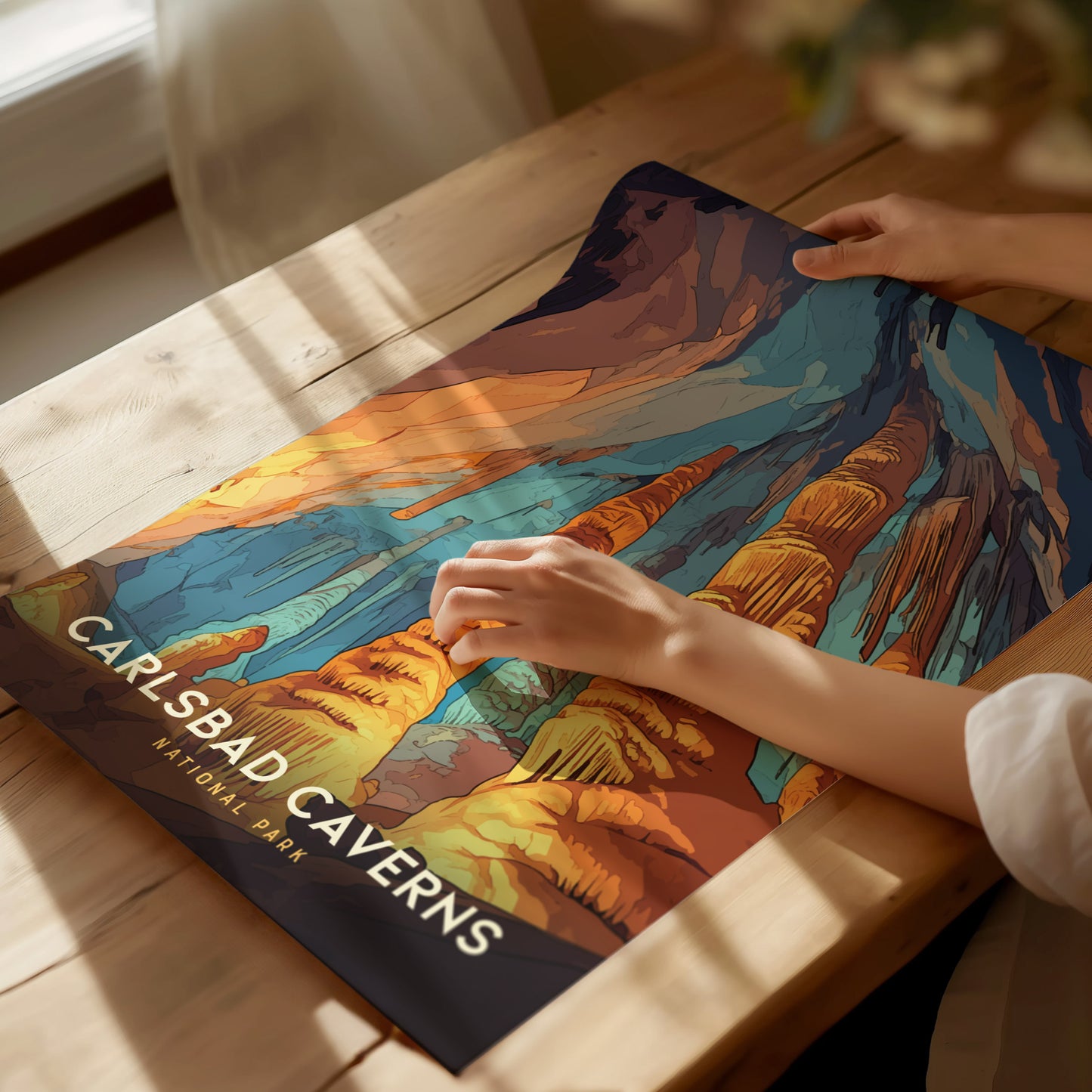 A person arranges a colorful Carlsbad Caverns National Park poster on a wooden table, featuring vibrant illustrations of rock formations and stalactites. Sunlight casts soft shadows across the scene.