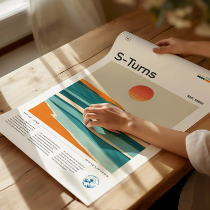 A person examines a colorful poster titled S-Turns, depicting abstract ocean waves and a setting sun, on a wooden table. Warm sunlight illuminates the scene.