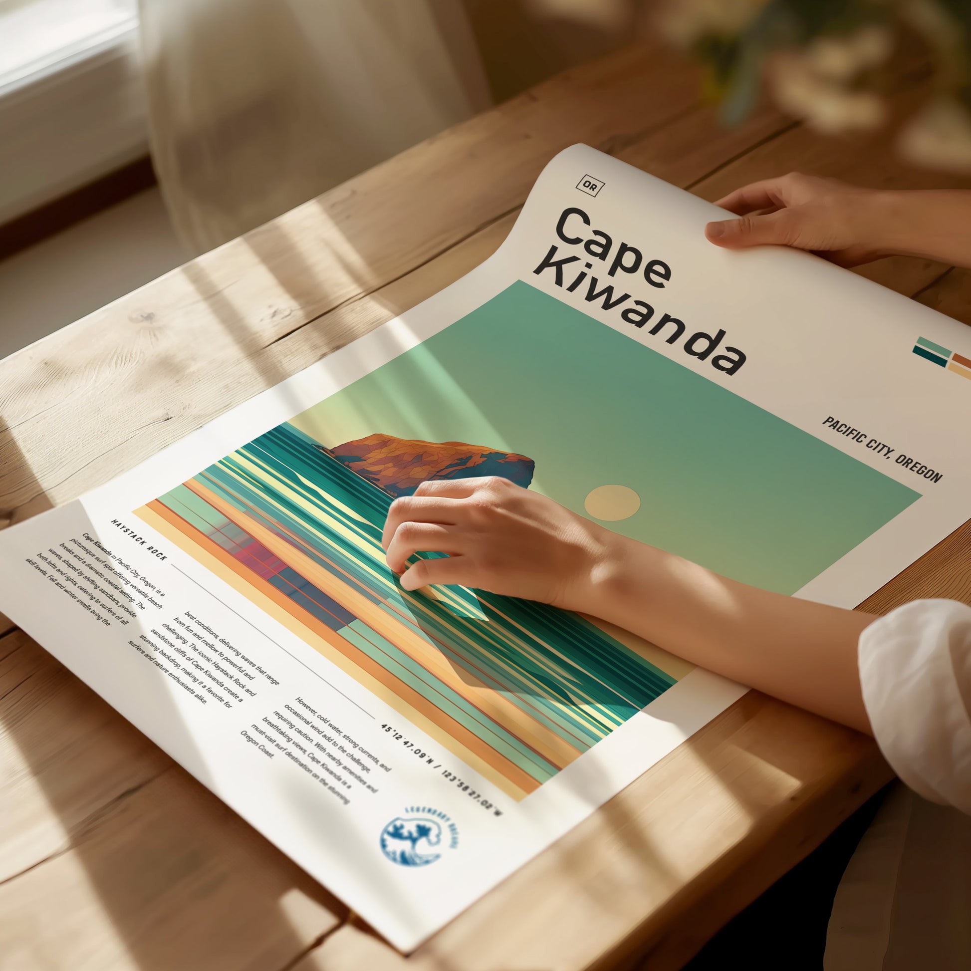 A person is placing a Cape Kiwanda poster on a wooden table. The poster features a stylized ocean scene with a rock formation and sun under a clear sky.