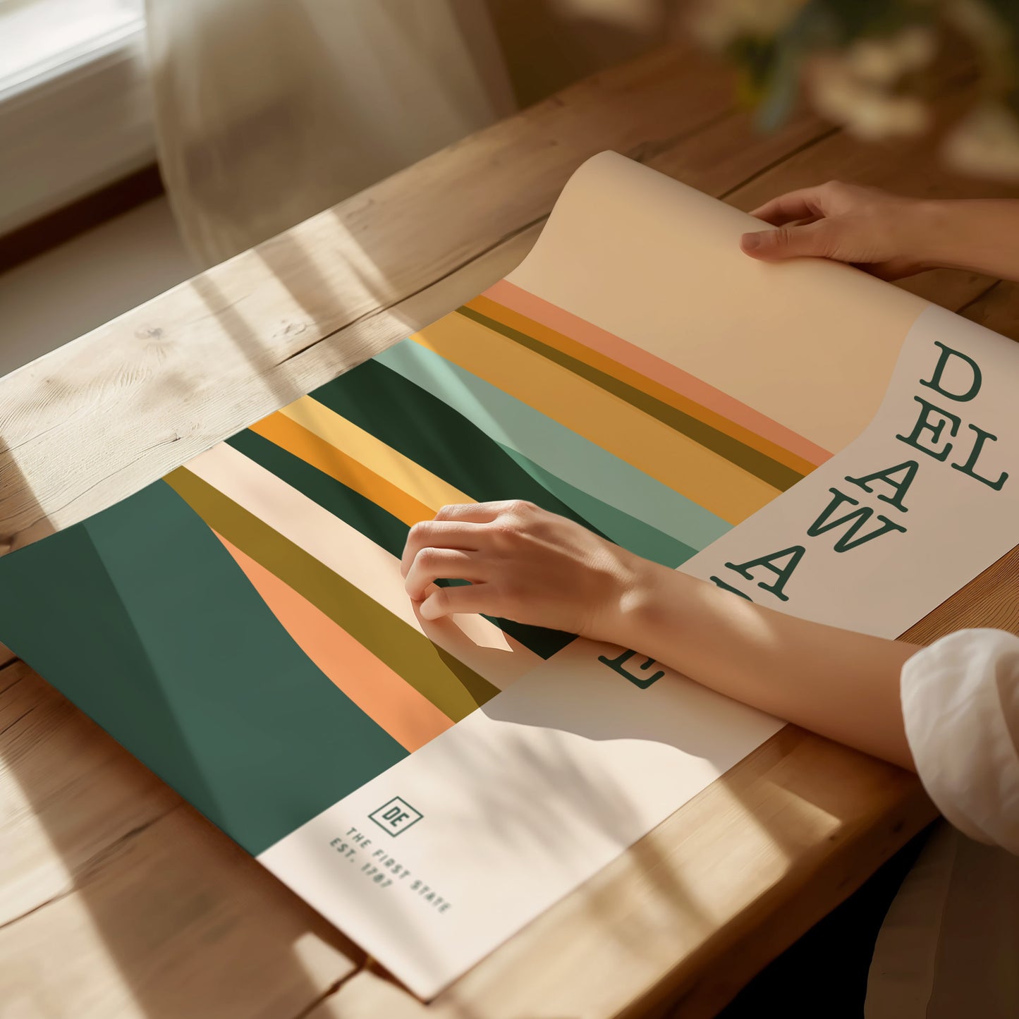 A person rolls out a minimalist Delaware poster featuring abstract, layered shapes in warm earthy tones on a wooden table. Sunlight casts gentle shadows across the scene.
