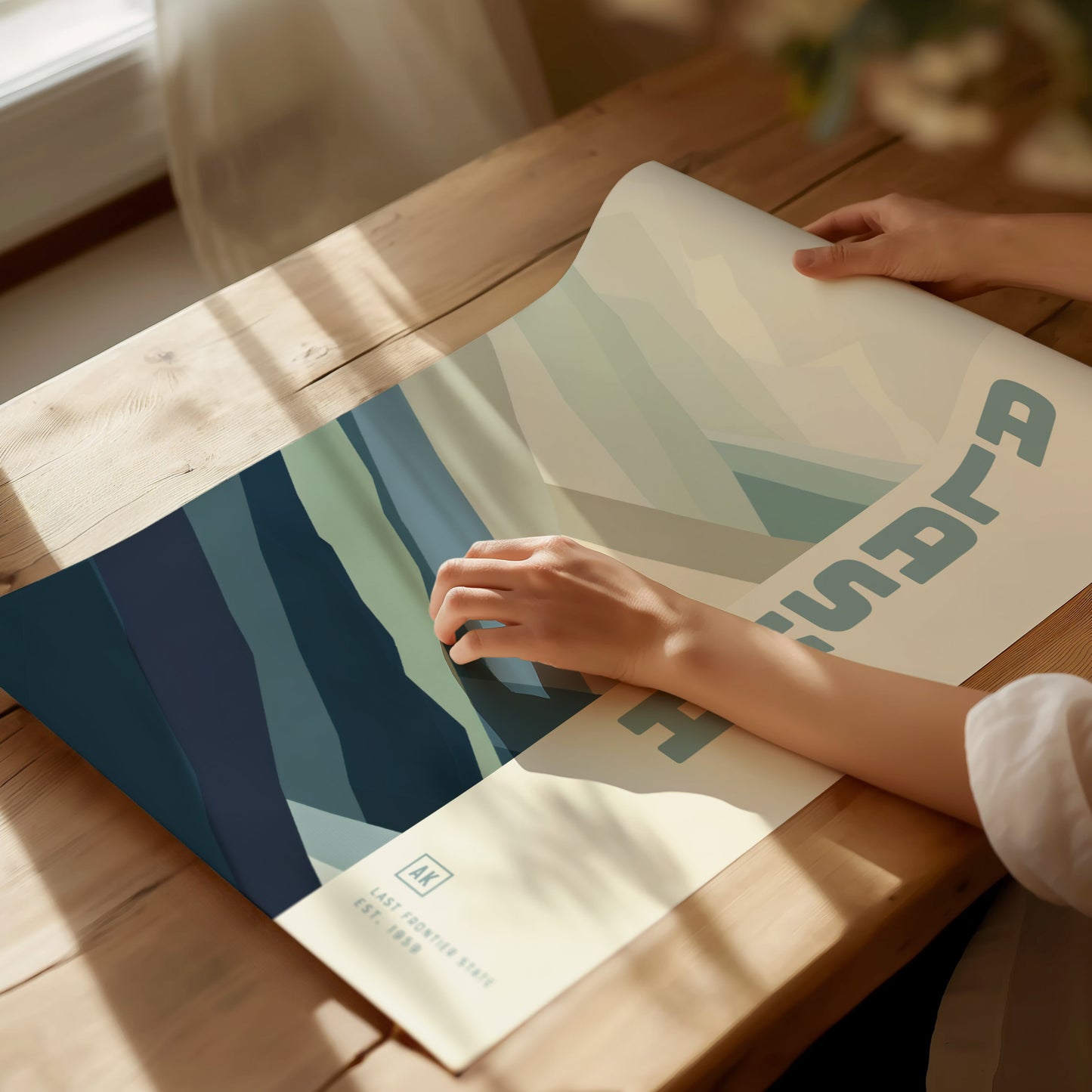 A person gently holds a minimalist poster featuring abstract blue and green shapes, with sunlight streaming onto a wooden table. The design includes text and artistic patterns.