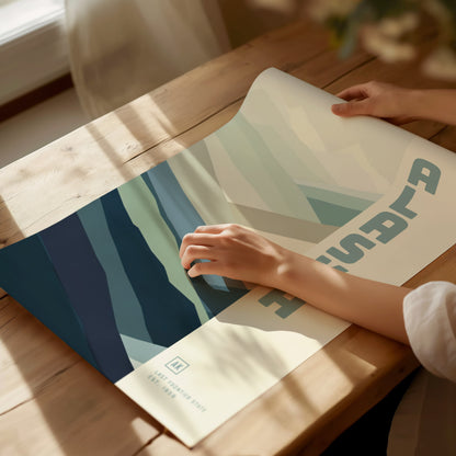 A person gently holds a minimalist poster featuring abstract blue and green shapes, with sunlight streaming onto a wooden table. The design includes text and artistic patterns.