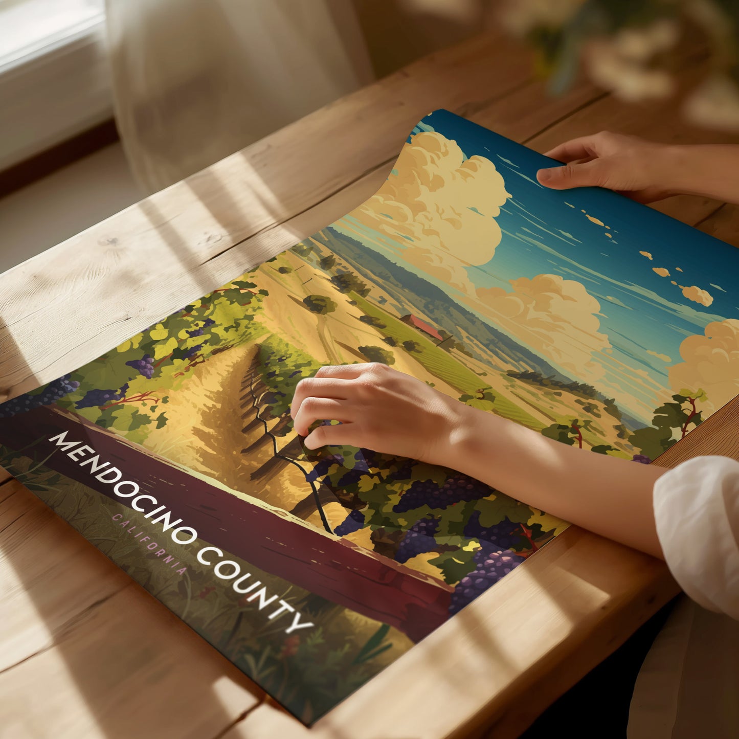 A person unrolls a vibrant Mendocino County poster on a wooden table, depicting a scenic vineyard landscape under a blue sky with fluffy clouds.