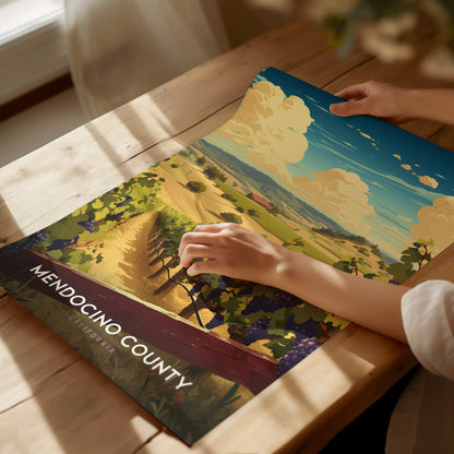 A person unrolls a vibrant Mendocino County poster on a wooden table, depicting a scenic vineyard landscape under a blue sky with fluffy clouds.
