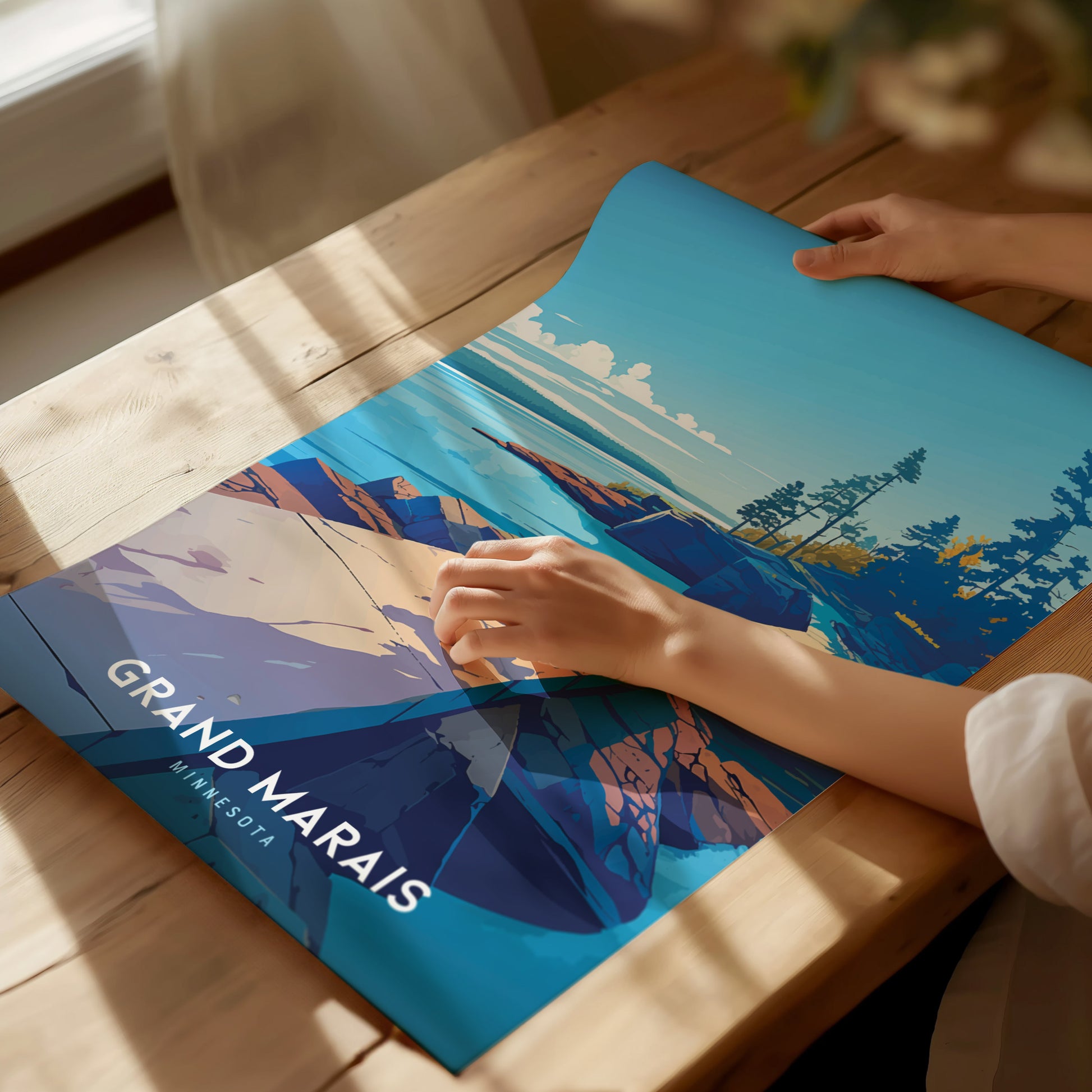 A person is unrolling a vibrant Grand Marais, Minnesota poster on a wooden table, showcasing a colorful landscape scene with trees and a lake under a blue sky.