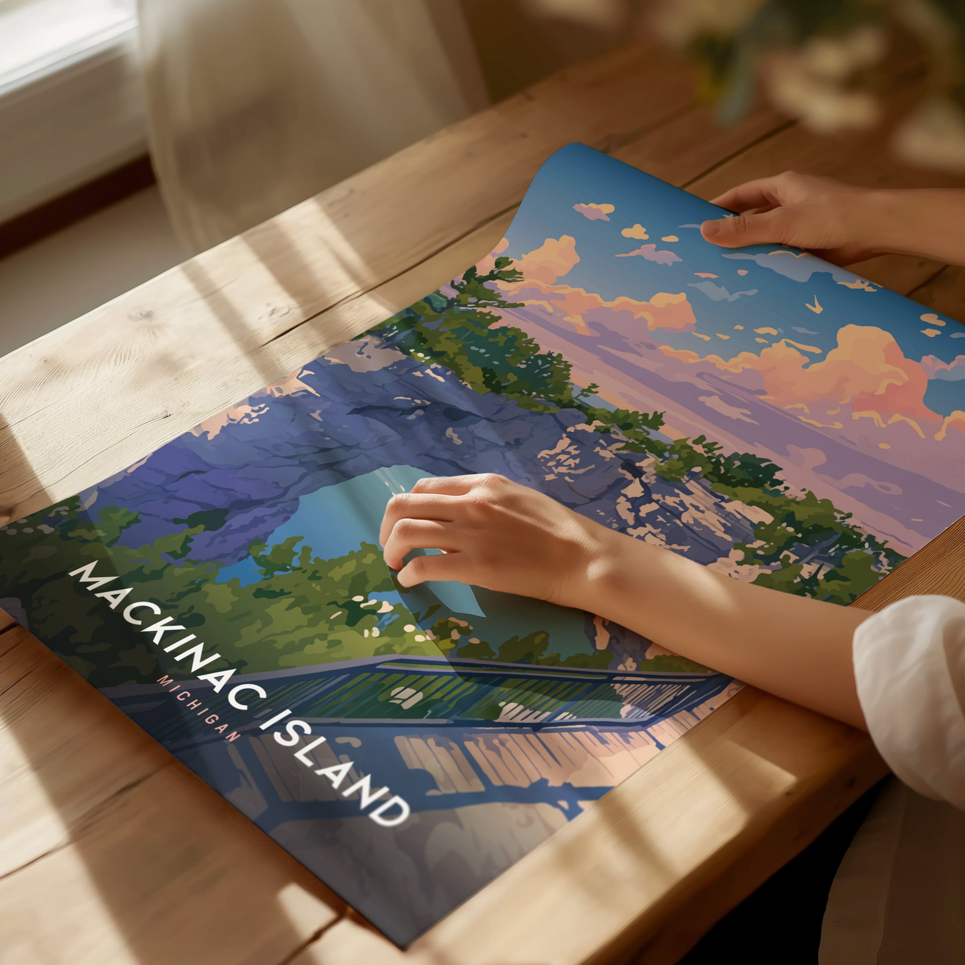 A person gently touches a colorful poster of Mackinac Island, showcasing a vibrant illustration of cliffs, trees, and a sunset sky. Sunlight casts soft shadows on the wooden table.