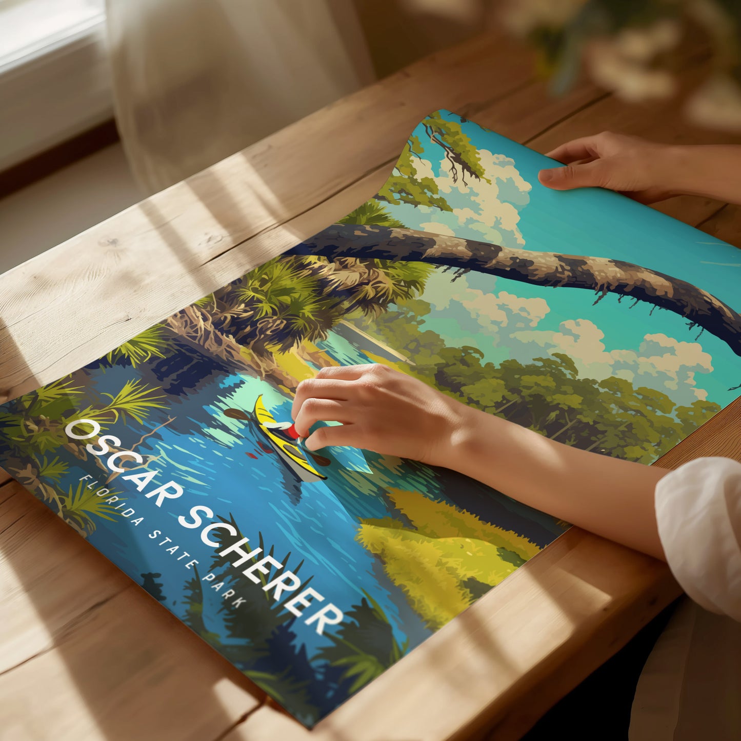 A person holds an Oscar Scherer State Park poster featuring vibrant nature scenery with water, trees, and a clear sky, placed on a wooden table. Sunlight casts soft shadows.