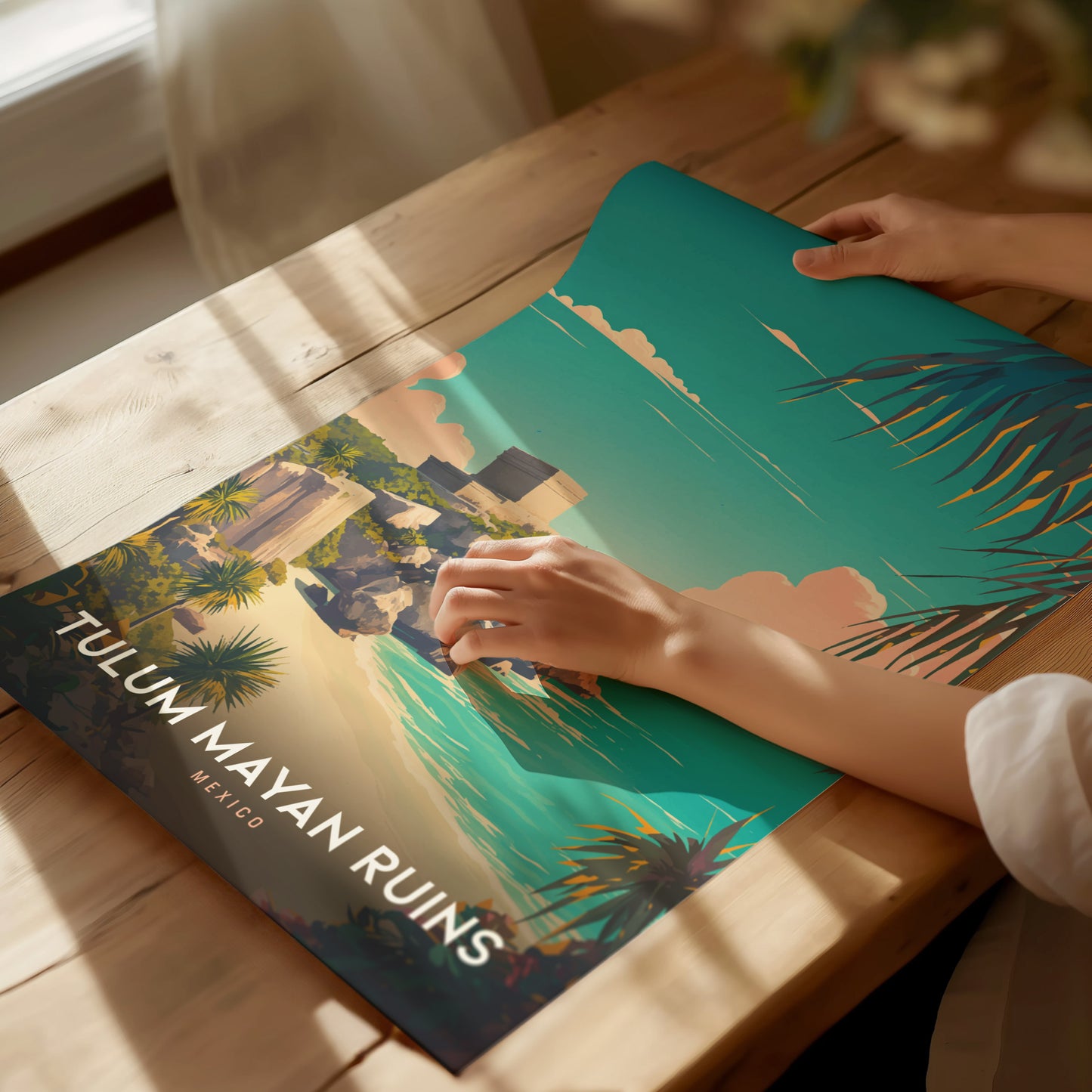 A person unrolls a vibrant Tulum Mayan Ruins poster on a sunlit wooden table, showcasing lush greenery, turquoise waters, and the iconic El Castillo temple.