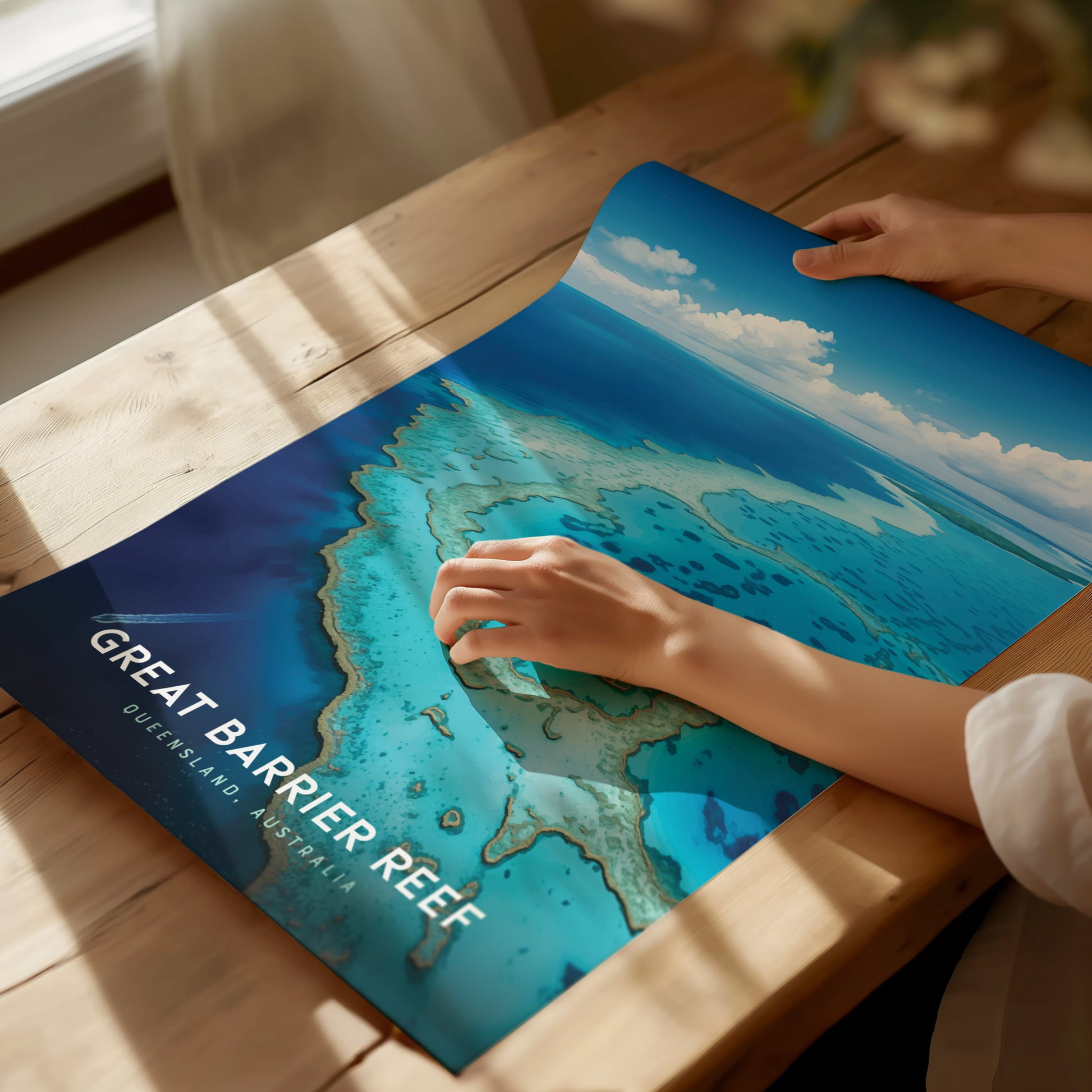 A person holds a vibrant Great Barrier Reef poster on a wooden table, showcasing turquoise waters and coral formations. Sunlight casts soft shadows across the surface.