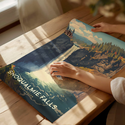 A person holds a vibrant Snoqualmie Falls poster on a wooden table, showcasing the waterfall amidst lush greenery. Sunlight casts gentle shadows across the scene.